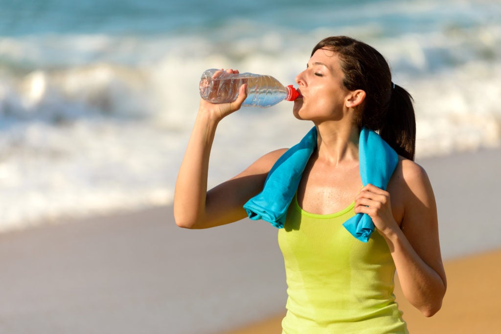 GIrl in yellow tanktop with blue towel around neck drinking water on a beach